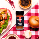 A bottle of Bravado Spice Garlic & Árbol Seasoning from Bravado Spice sits on a red checkered cloth beside a sandwich, onion, roasted garlic, black pepper, red chilies, and small bowls of spices—perfect for adding a Tex-Mex spice twist to your favorite dishes.