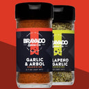Two spice jars from Bravado Spice Co are displayed: one is the Bravado Spice Co Garlic & Árbol Seasoning, featuring a bold Tex-Mex blend with vibrant red spices, and the other is Jalapeño Garlic Seasoning filled with green spices, both set against a striking red geometric background.