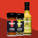 Three Bravado Spice Co. items—a bottle of Bravado Spice Co. Garlic & Árbol Seasoning, Jalapeno & Garlic Seasoning, and Creamy Herb & Jalapeno Hot Sauce—are showcased against a vibrant red geometric backdrop, perfect for anyone craving the bold, authentic flavors of Tex-Mex seasoning.