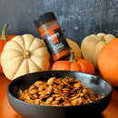 A black bowl of seasoned pumpkin seeds sits in front of assorted pumpkins, with a jar of Bravado Spice Co Garlic & Árbol Seasoning resting on a white pumpkin.
