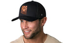 A smiling man with a trimmed beard wears the Haka Hat Arizona Flag Trucker Hat Leather— a black snapback by Haka Hat featuring an orange Arizona state outline and star— and a beige shirt, against a white background.