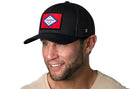 A man with a trimmed beard wears the black Haka Hat Arkansas Flag Trucker Hat, a mesh-back snapback featuring a state flag patch. He smiles in a beige shirt against a white background.