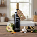 A bottle of Georgetown Olive Oil Co. ARTICHOKE & GARLIC Infused | High Polyphenols Extra Virgin Olive Oil stands on a kitchen counter, accompanied by a sliced artichoke, a garlic bulb, and a bowl of mixed olives. A product tag hangs from the bottle neck while blurred kitchen shelves fill the background.