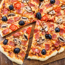 A sliced pizza with black olives, mushrooms, cherry tomatoes, ham, and melted cheese sits on a wooden board, one slice slightly pulled out and finished with SALTOPIA Infused Sea Salts’ Saltopia Hot Honey for a touch of irresistible sweet heat.