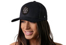 A woman with long brown hair smiles while wearing the Haka Hat Austin Trucker Hat | Black ATX X Snapback, a black mesh snapback by Haka Hat featuring a circular ATX embroidered patch. The background is white.