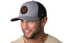 A smiling man with a short beard wears the Haka Hat Austin Trucker Hat, a gray-black snapback featuring an orange ATX X embroidered patch. He pairs it with a light beige shirt against a white background.