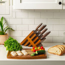 The Messermeister Avanta Pakkawood Knife Block Set - 10 Piece sits on a white kitchen counter near a cutting board with lettuce, cherry tomatoes, cucumber, scallops, and sliced bread. A potted plant and tiled backsplash are in the background.