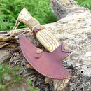 The Texan Knives Custom Hand Engraved Ax Pizza Cutter with a J2 steel blade, wooden handle, and brown leather sheath rests on a weathered log amid green foliage.