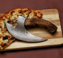 The Texan Knives Axe Pizza Cutter—Custom Hand-Engraved J2 Steel Blade with Wooden Handle—rests on a wooden cutting board beside a sliced pizza.