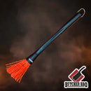 The Butcher BBQ Mop Brush features a long black handle, bright red silicone bristles, and a metal hook—ideal for applying sauce. Set against a smoky backdrop with the Butcher BBQ logo in the corner.
