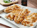 Four BBQ chicken flatbreads, topped with melted cheese, chopped red onions, and cilantro, rest on a baking tray. Terrapin Ridge Farms Sweet Heat Grill & Wing Squeeze and a cutting board with chopped onions are in the background.
