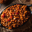 Close-up of baked beans in a cast iron skillet with red pepper and onion, enhanced by SALTOPIA Infused Sea Salts' BBQ Salt: MEAT HEAD, on a rustic wooden surface with burlap underneath.