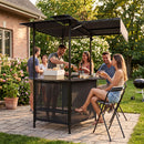 Five people enjoy drinks in the sun under the DirectDeals Grill Gazebo with Shelves, Hooks & Double Roof Canopy on a stone patio, surrounded by plants and with a grill nearby.