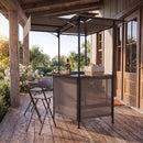 The DirectDeals Grill Gazebo with Shelves, Hooks & Double Roof Canopy sits on a wooden porch with a folding chair, drinks, and bread basket, surrounded by greenery and flowers in bright daylight.