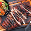 Barbecued ribs sliced on a wooden board with grilled peppers and zucchini, a steak knife alongside, are finished with SALTOPIA Infused Sea Salts' Applewood Smoked Salt: GREAT OUTDOORS for a rich, smoky flavor boost.