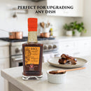 A bottle of Taycte Aged Balsamic Steak Sauce sits on the kitchen counter beside a plate of ribs and a small bowl of sauce. Text above reads, Perfect for upgrading any dish. Kitchen appliances and utensils in the background emphasize the rich flavor of Taycte’s steak sauce, expertly crafted with 25-30 year Balsamic Vinegar of Modena.