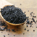 A wooden spoon filled with SALTOPIA Infused Sea Salts' Saltopia Black Lava Salt rests on a wooden surface, with some of the coarse finishing salt granules spilling onto the table.