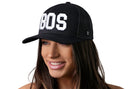 A woman with long brown hair smiles while wearing the Haka Hat Boston White Block Trucker Hat, a black Massachusetts snapback by Haka Hat with bold BOS lettering. The background is white.