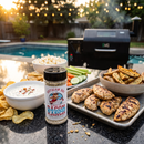 A container of BuffalowBBQ Bacon Ranch BBQ Rub stands on a table next to a tray of grilled chicken, bowls of dip and potato wedges, and a platter of vegetables.