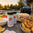 Two jars of BuffalowBBQ Bacon Ranch BBQ Rub on an outdoor table beside grilled chicken, potato wedges, popcorn, and dip, with a grill and swimming pool in the background.