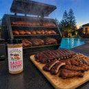 A bottle of BuffalowBBQ Sassy Swine BBQ Rub stands next to a wooden cutting board piled with smoked ribs, chicken drumsticks, and pulled pork, with a smoking grill in the background by a swimming pool.