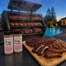 Two bottles of BuffalowBBQ Sassy Swine BBQ Rub sit on a table next to a wooden cutting board piled with ribs, pulled pork, and chicken, with a large smoker filled with meat in the background by a swimming pool.