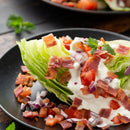 A wedge of iceberg lettuce topped with creamy dressing, diced tomatoes, red onions, and crispy bacon, finished with SALTOPIA Infused Sea Salts’ HOWDY DO Bacon & Ranch Seasoning, served on a black plate with a mint garnish.
