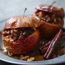 Two baked apples stuffed with nuts and spices sit on a plate, topped with SALTOPIA Cinnamon Sugar: CRACKLIN' HEIGHTS 16oz, adding a warm, sweet finish from SALTOPIA Infused Sea Salts. Cinnamon sticks and syrup complete the presentation.