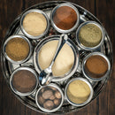 The Spice Wheel Baking Spice Wheel by Spice Wheel serves as a stylish spice organizer, featuring eight small containers filled with assorted ground baking spices and whole nutmeg, plus a central compartment for granulated sugar with an included spoon—all beautifully arranged on a dark wooden surface.