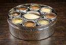 The Spice Wheel Baking Spice Wheel by Spice Wheel is a round stainless steel spice organizer featuring nine compartments filled with assorted ground baking spices and comes with a small spoon, displayed on a wooden surface.
