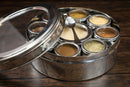 The Spice Wheel Baking Spice Wheel by Spice Wheel is a round metal spice box with a clear lid, featuring seven small containers filled with assorted ground spices and a small metal spoon, displayed on a wooden surface.