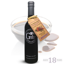 A bottle of Georgetown Olive Oil Co. 18 Year Aged Balsamic Vinegar Reduction stands before a bowl of balsamic, its tag highlighting that it’s aged 18 years in the tradition of Modena. Some spilled vinegar and the text “AGED 18 YEARS” appear in the lower right corner.