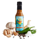 A bottle of Culture Hot Sauce Bar Sauce from Culture Hot Sauce rests among fresh garlic bulbs, jalapeño peppers, salt, and peppercorns. Its label reads “Bar Sauce” and features an illustrated beach scene.