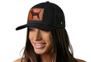 A smiling woman with long brown hair wears a black Haka Hat Beagle Trucker Hat Leather Dog Mom Snapback featuring a dog silhouette patch, against a plain white background.