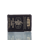 A black-wrapped bar of The Immaculate Beard Natural Beard Wash Bar is displayed upright on a reflective surface, its label showcasing product details and ingredients. This natural beard care essential also functions as an activated charcoal shampoo, providing a convenient beard shampoo bar for deep cleansing and lasting freshness.
