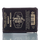 A bar from The Immaculate Beard, included in the Immaculate Premium Beard Grooming Gift Set For Men Complete Care Kit, is wrapped in dark packaging with white text. The label highlights its herba-mint, lemon peel, lavender, and sea salt scents. Ideal for any beard grooming kit, the bar is displayed on a reflective surface.