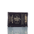 A rectangular bar of The Immaculate Beard’s Immaculate Beard Natural Wash Bar For Men Deep Clean, a natural beard wash and shampoo infused with activated charcoal, wrapped in dark packaging featuring white and yellow text, is displayed on a reflective surface.