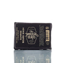 A bar of The Immaculate Beard Natural Beard Wash Bar, a beard shampoo designed for natural beard care, is wrapped in dark paper featuring white and gold text and sits atop a reflective surface. Packaging displays the ingredients and product details clearly.