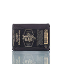 A bar of The Immaculate Beard’s Natural Beard Wash Bar, wrapped in black paper with gold and white lettering and reflecting on a white surface. This handmade beard shampoo bar is perfect for natural beard care and features a refreshing citrus, vanilla musk scent.