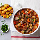 A white pot of beef stew with beef, carrots, mushrooms, and herbs sits on a cloth beside roasted potatoes seasoned with SALTOPIA BLT Salt: BACON. LETTUCE. TOMATO. from SALTOPIA Infused Sea Salts and a dish of chopped parsley.
