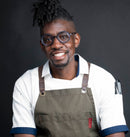 A smiling man with glasses and styled hair stands against a plain dark background, wearing a white shirt, green apron, and pen in pocket—ready to share the bold aromas of Middle Eastern flavors with his signature World Spice Kaffe Baharat blend from World Spice.