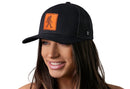 A woman with long brown hair is smiling and wearing a Haka Hat Bigfoot Leather Trucker Hat, a black Sasquatch snapback by Haka Hat, featuring an orange patch with a Bigfoot silhouette. The background is white.