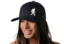 A woman with long brown hair smiles while wearing a Haka Hat Bigfoot Trucker Hat, a black Sasquatch Snapback featuring a white silhouette of Bigfoot on the front. The background is plain white.