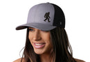 A woman with long brown hair smiles while wearing the Haka Hat Bigfoot Trucker Hat, a gray-black Sasquatch snapback by Haka Hat featuring a Sasquatch silhouette on the front panel. The background is plain white.