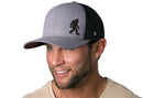 A smiling man in a beige shirt and light facial hair wears the Haka Hat Bigfoot Trucker Hat—a gray and black snapback by Haka Hat featuring a Sasquatch silhouette on the front panel.