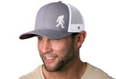 A man with trimmed facial hair wears the Haka Hat Bigfoot Trucker Hat, a gray and white Sasquatch snapback by Haka Hat, paired with a beige T-shirt, smiling and looking slightly to the side.