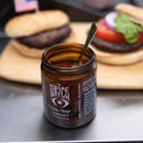 An open jar from the Spice Witch Chili Crisp Box of 3 with a spoon inside sits in front of two hamburgers on a wooden tray, one topped with tomato, onion, and lettuce.