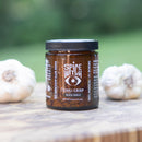 A box of 2 Spice Witch Chili Crisp jars rests on a wooden surface beside two whole garlic bulbs, with a blurred green background.