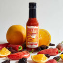 A bottle of Culture Hot Sauce Blindsided by Culture Hot Sauce—a smoky citrus hot sauce—is surrounded by whole and sliced oranges, red chili peppers, and dried chilies on a white background.