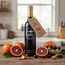 A bottle of Georgetown Olive Oil Co Blood Orange Fused | High Polyphenols Extra Virgin Olive Oil from Georgetown Olive Oil Co. sits on a kitchen counter, surrounded by whole and sliced blood oranges and an assortment of black and green olives. The bottle features a gold cap and a label with a hanging tag, emphasizing its high polyphenols extra virgin olive oil content for enhanced flavor and health benefits.